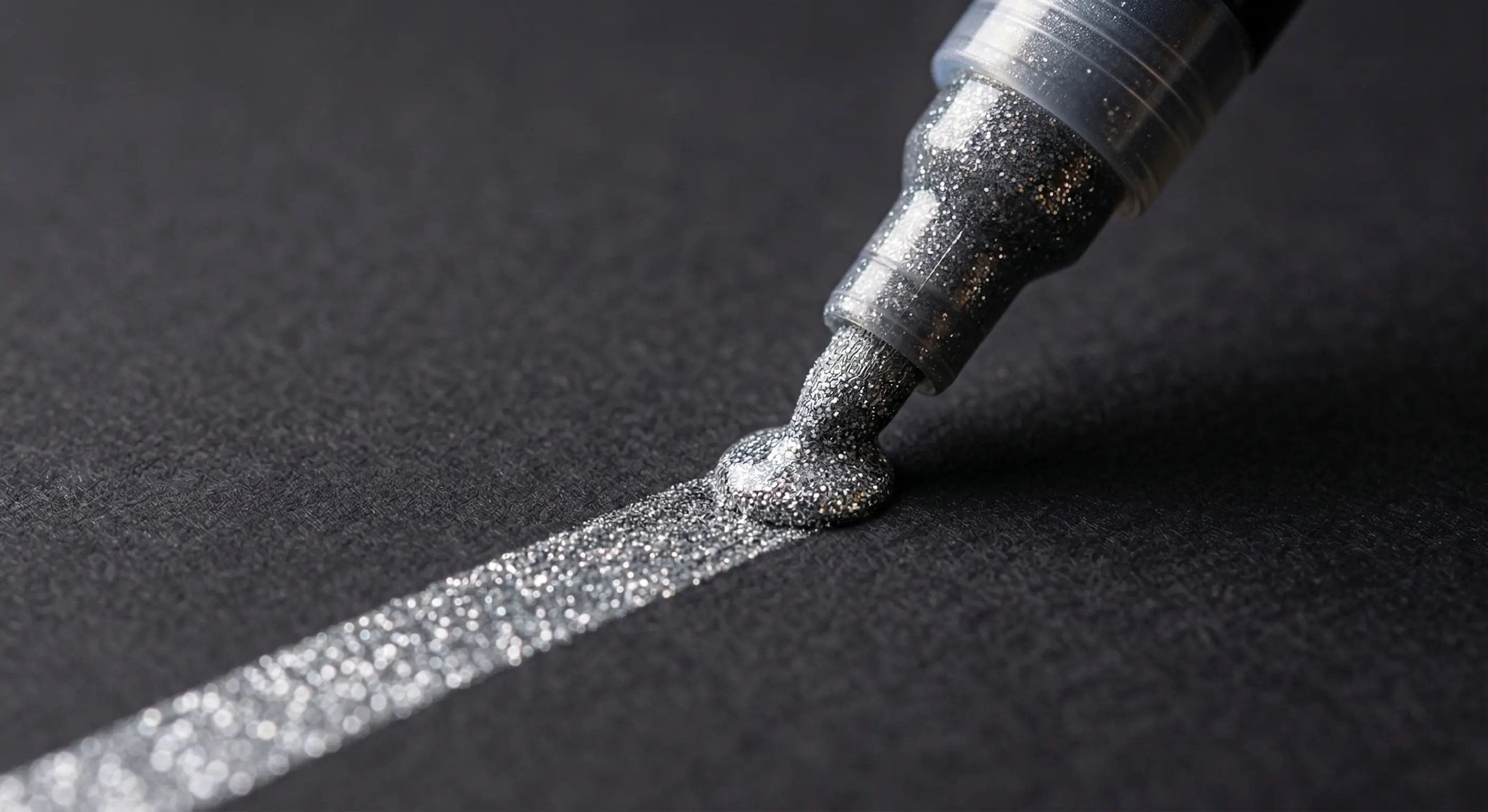 A macro close-up of a pen nib drawing a shimmering, opaque rainbow-colored glitter line on dark black cardstock.