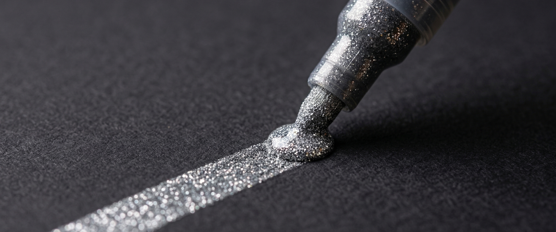A macro close-up of a pen nib drawing a shimmering, opaque rainbow-colored glitter line on dark black cardstock.